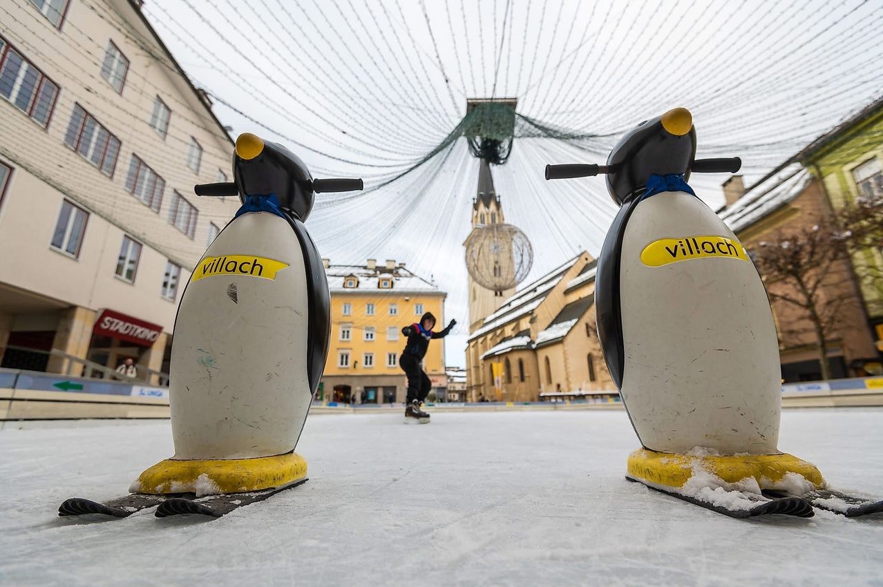 Ein Skater fährt an Pinguinskulpturen auf einer Eisbahn vorbei, mit Gebäuden und einer Kirche im Hintergrund.