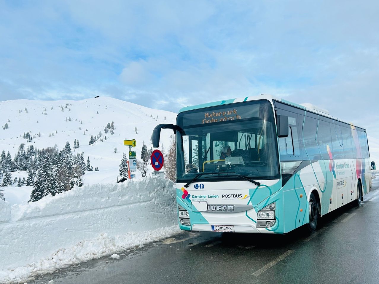Ein blau-weißer Bus fährt auf einer verschneiten Straße mit Bergen im Hintergrund. Der Bus hat ein Zielschild mit der Aufschrift 'Naturpark Nobertach'.