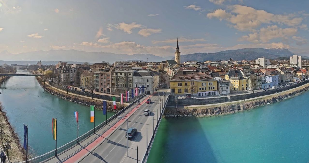 Eine Stadt mit Gebäuden, Flaggen, einer Brücke, einem Fluss und Bergen in der Ferne. Autos fahren auf der Brücke.