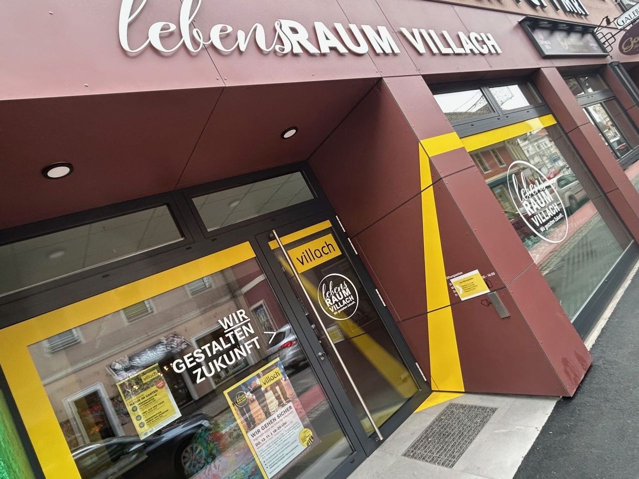 Der Eingang eines Geschäfts namens 'Lebensraum Villach'. Es hat eine Glastür mit gelben Rändern und einem gelben Streifen. Das Schild des Geschäfts ist prominent oben angebracht. Die Tür hat ein gelbes Etikett mit dem Text 'Lebensraum Villach'. Ein Plakat mit dem Text 'Wir gestalten Zukunft' ist an der Glastür befestigt.