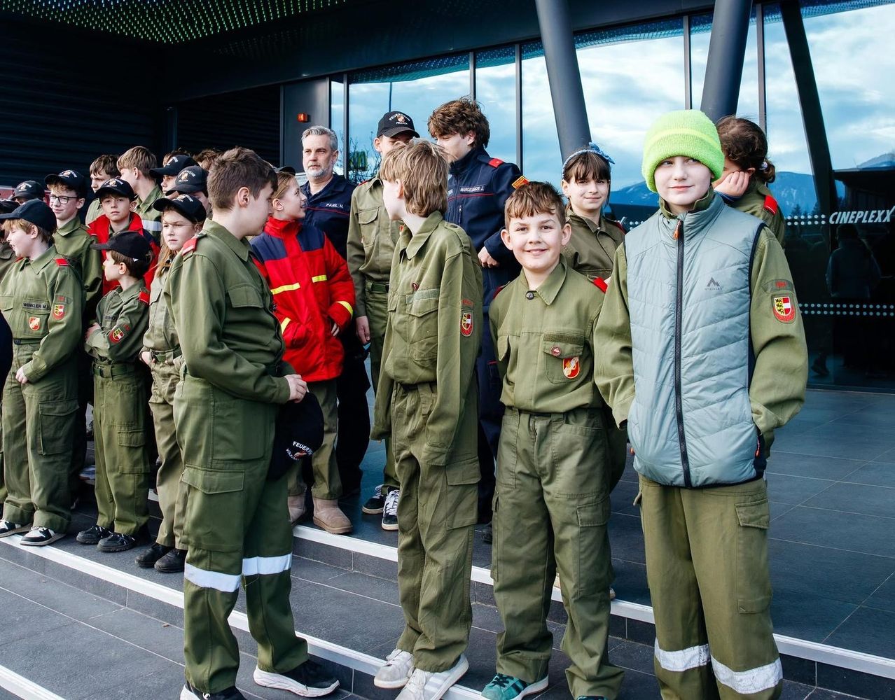 Eine Gruppe von Kindern in grünen Uniformen steht in einer Reihe, mit einem erwachsenen Mann in einem roten Jackett hinter ihnen. Sie scheinen Teil einer Parade oder einer besonderen Veranstaltung zu sein.