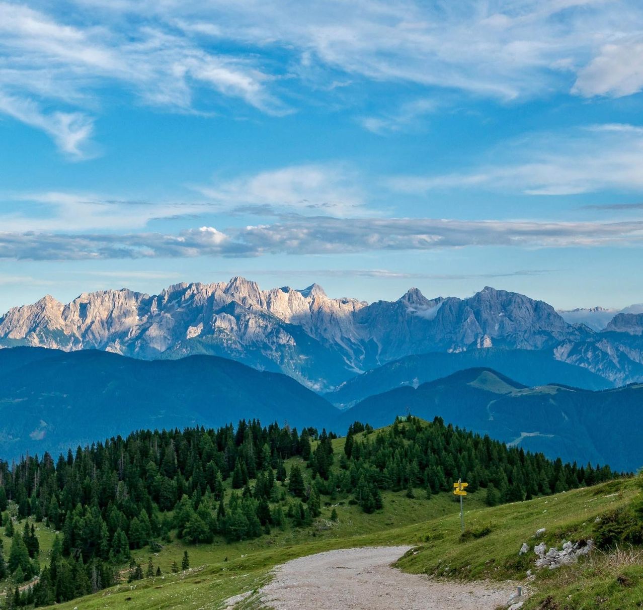 Eine malerische Aussicht auf eine Bergkette mit einem unbefestigten Weg im Vordergrund. Der Himmel ist blau mit verstreuten Wolken.
