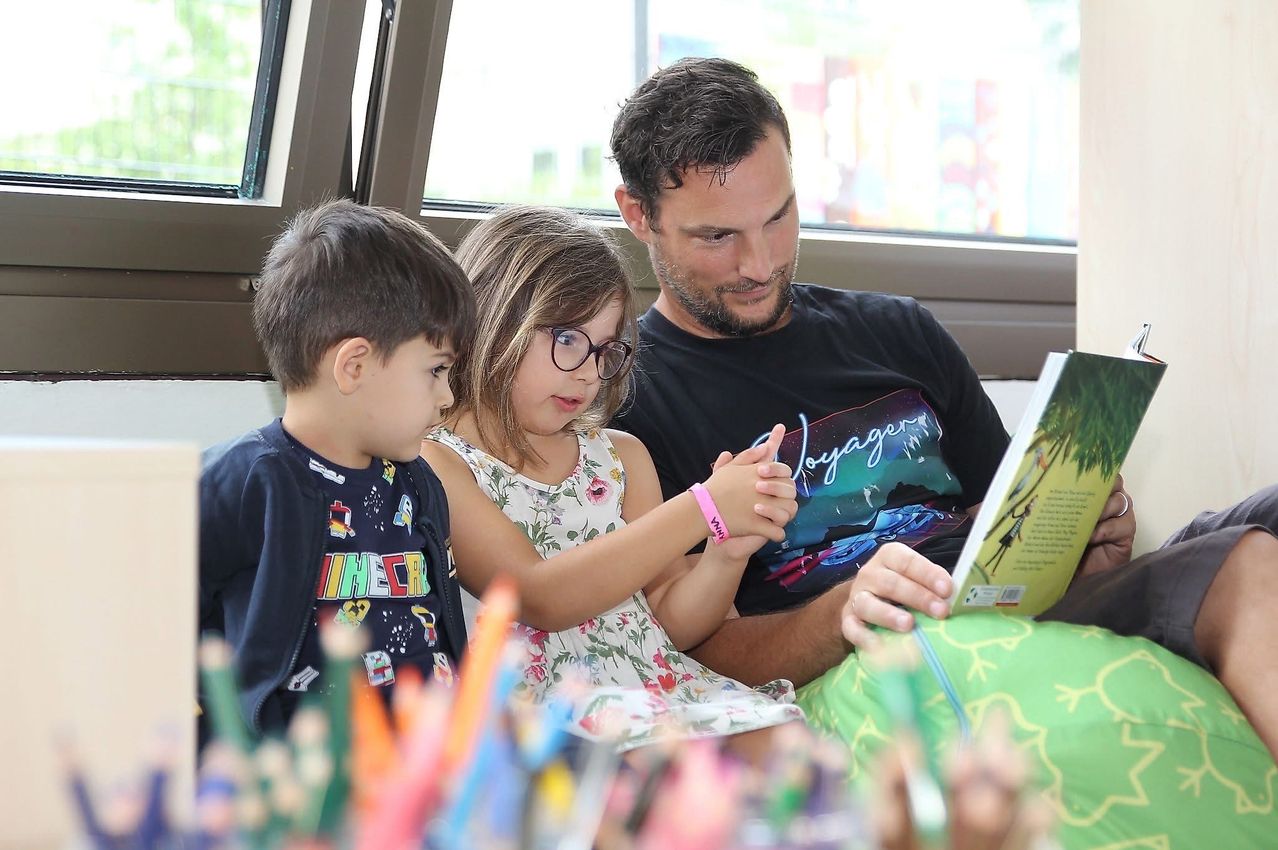 Ein Mann liest einem Mädchen und einem Jungen ein Buch vor, die mit farbenfrohen Stiften auf einem Tisch sitzen. Der Mann trägt ein schwarzes T-Shirt.
