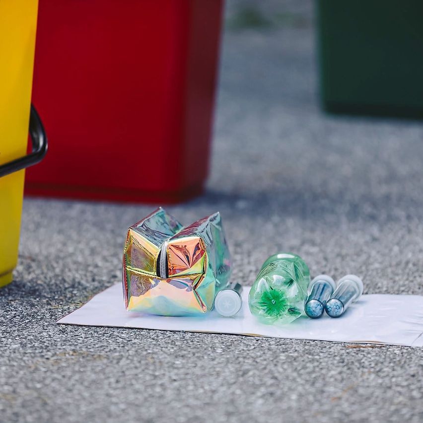 Eine farbenfrohe Tasche mit Reißverschluss und einige kleine Behälter liegen auf einem Stück Papier am Boden neben Mülleimern.