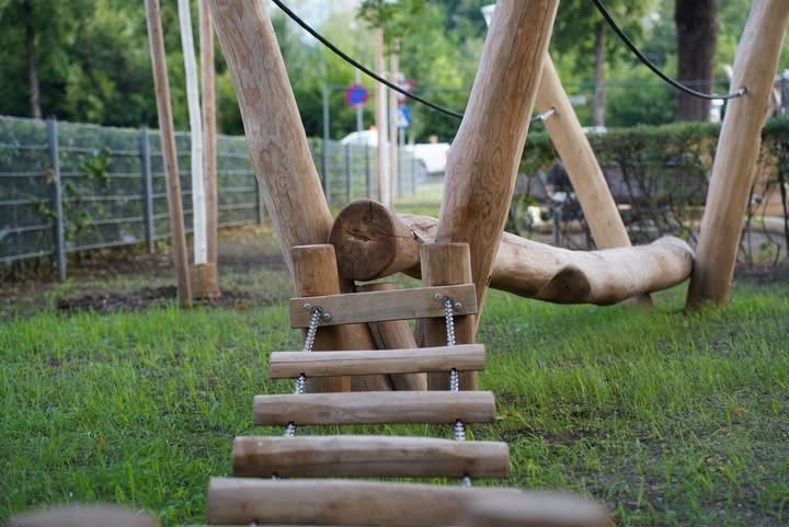 Eine Holzkletterstruktur mit einer Leiter und Baumstämmen in einem Grasbereich. Bäume und ein Zaun sind im Hintergrund.