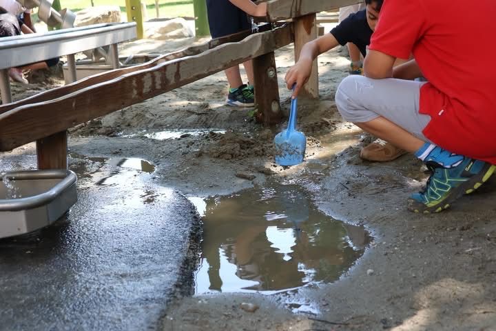 Kinder spielen in einem Sandkasten mit einem blauen Schaufel, ein Junge im roten Shirt hockt sich neben einer Pfütze.