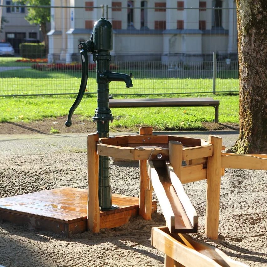 Eine grüne, alte Pumpe steht auf einer Holzplattform in einem Park mit einer Holzrutsche und einem Baumstamm in der Nähe.