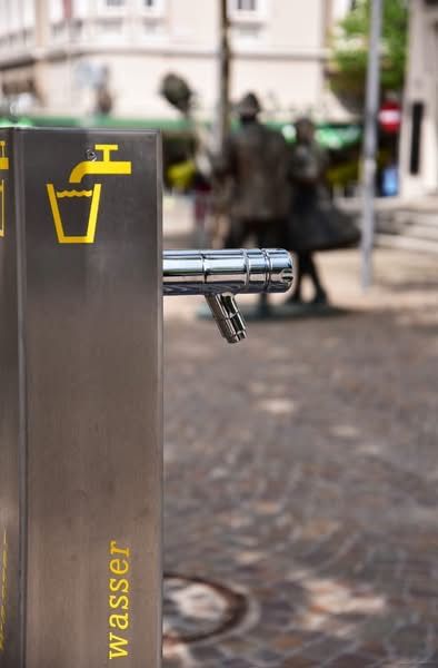 Eine Edelstahl-Wasserbrunnen mit einem gelben Wasserhahn und dem Wort Wasser darauf. Im Hintergrund ist ein verschwommenes Bild einer Statue und eines Gebäudes zu sehen.