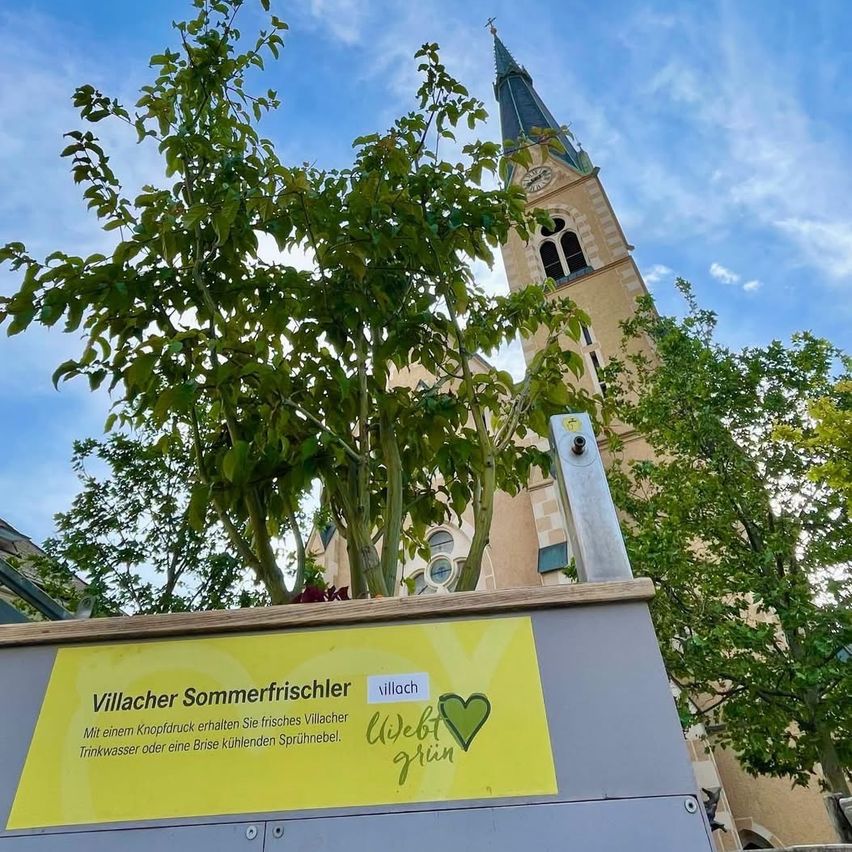Ein gelbes Schild mit grünem Text ist an einem Metallpfosten vor einer Kirche befestigt. Das Schild trägt die Aufschrift 'Villacher Sommerfrischer' mit einem Herzsymbol und dem Wort 'Web'. Die Kirche hat einen Glockenturm und einen Kirchturm. Vor dem Schild steht ein Baum.