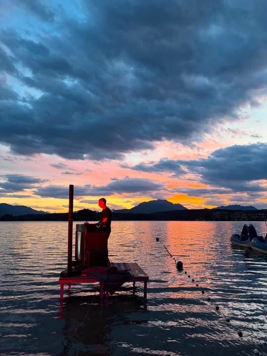 Ein Mann steht auf einer kleinen Plattform mitten im See, mit Bergen im Hintergrund und einem Sonnenuntergang am Himmel.