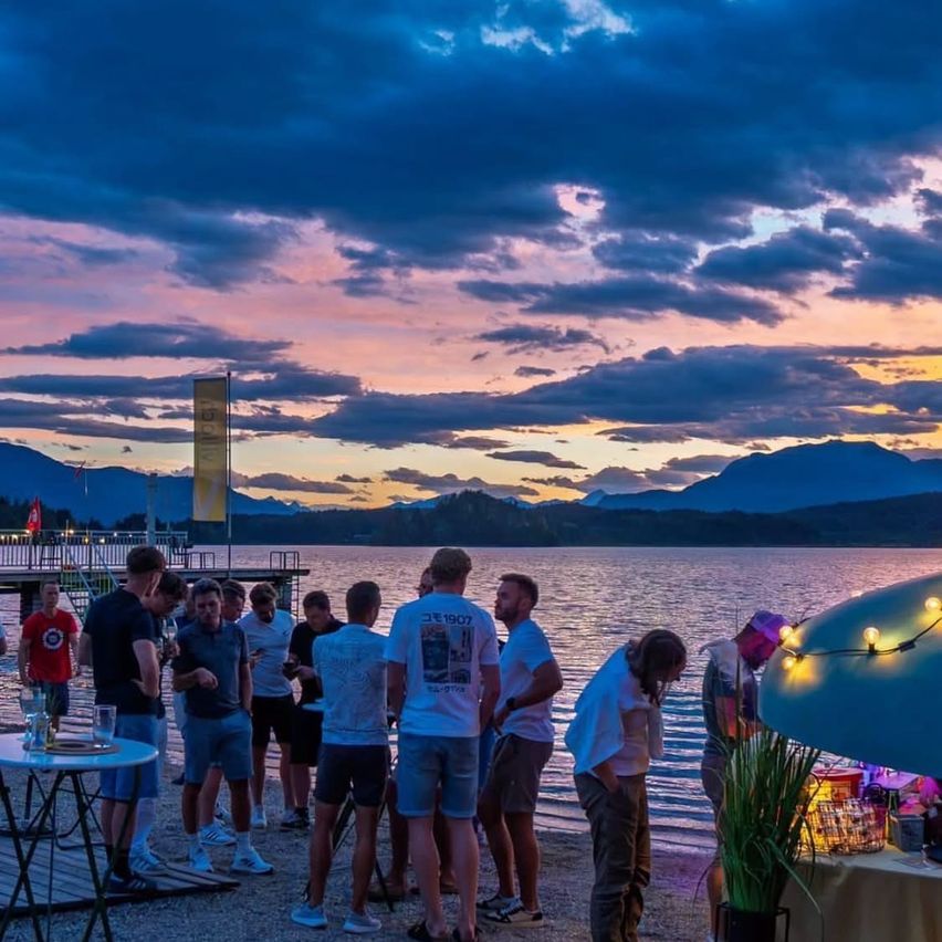 Eine Gruppe von Menschen steht am See und plaudert, während sie den Sonnenuntergang mit Bergen im Hintergrund genießt.