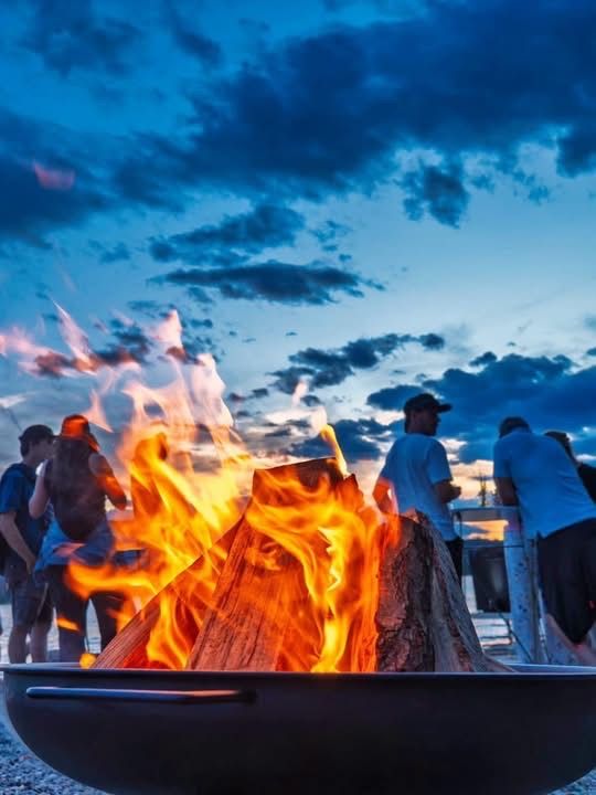 Eine Gruppe von Menschen versammelt sich um einen großen Feuerkorb und genießt die Wärme und das Licht der Flammen unter einem bewölkten Himmel.