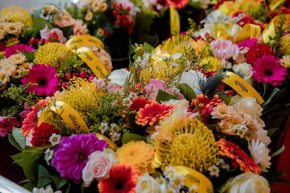 Ein farbenfroher Blumenstrauß mit gelben Bändern, einschließlich Rosen, Gänseblümchen und anderen bunten Blumen, in einem kreisförmigen Muster angeordnet.