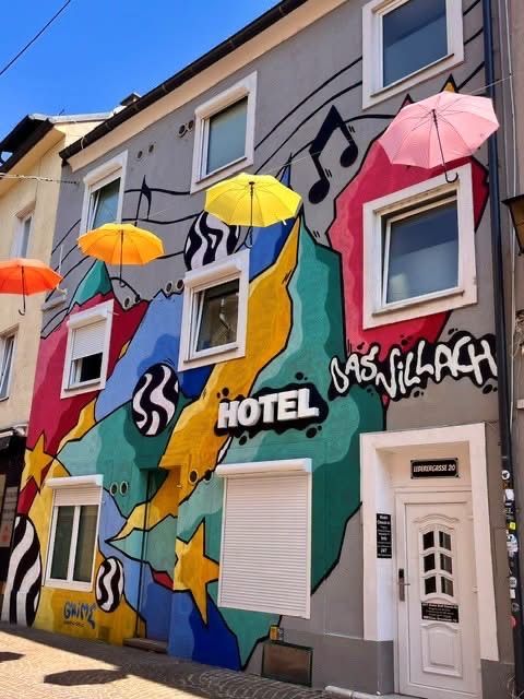 Ein farbenfrohes Gebäude mit einer Wandmalerei, die Musiknoten, Sterne und Regenschirme zeigt. Das Wort 'Hotel' ist prominent angebracht, und es gibt mehrere Fenster und eine weiße Tür.