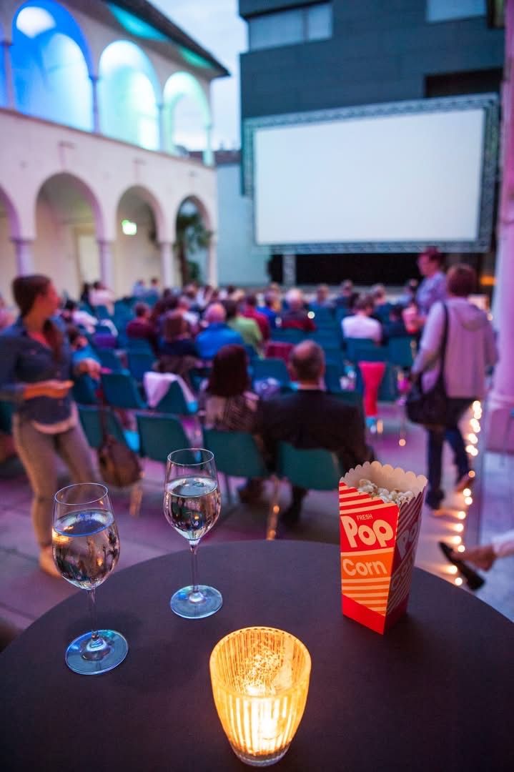 Ein Kinosaal mit einer großen Leinwand, auf der ein Film läuft, eine Menschenmenge in Stühlen sitzend und ein Tisch mit zwei Gläsern Wasser und einer Packung Popcorn.