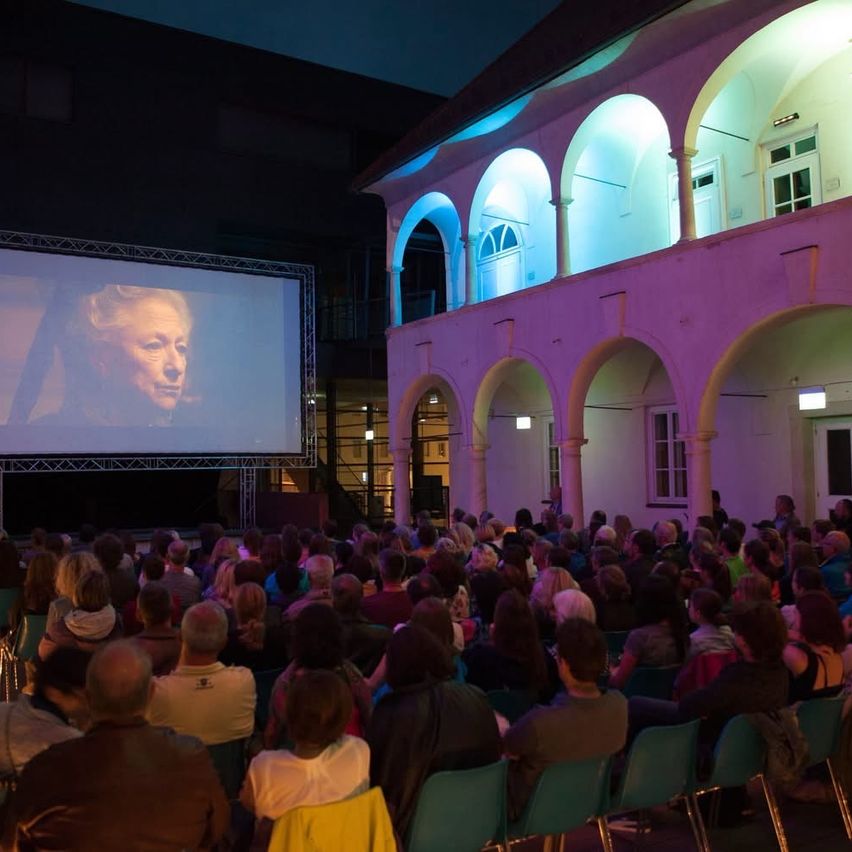 Eine große Menschenmenge sitzt auf Stühlen und schaut sich einen Film an, der auf eine Leinwand vor einem Gebäude mit gewölbten Fenstern projiziert wird, das von lila Lichtern beleuchtet ist.