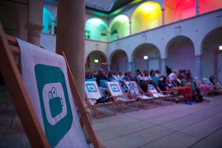 Eine Gruppe von Menschen sitzt auf Liegestühlen in einem Außenbereich, möglicherweise für eine Filmvorführung. Die Stühle sind in Reihen angeordnet und ein Banner mit einem Filmprojektor-Logo ist links platziert. Der Bereich ist von farbigen Lichtern beleuchtet.