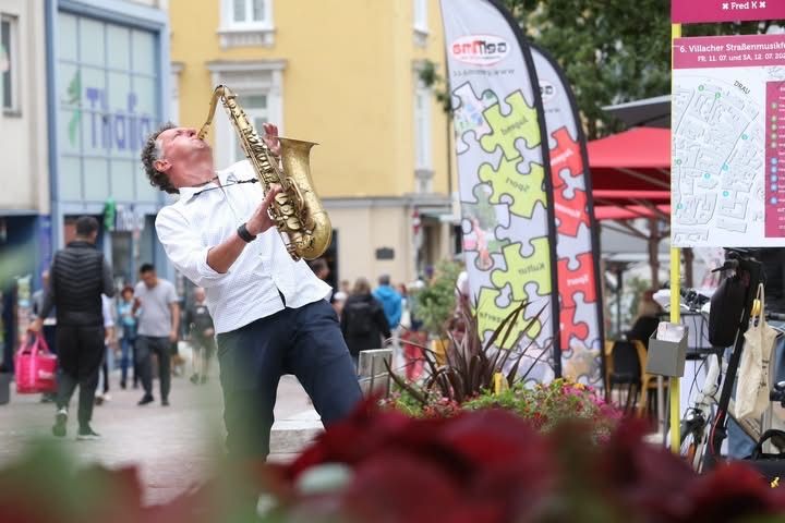 Ein Mann spielt Saxophon auf einer Straße, mit vorbeigehenden Leuten und einem roten Regenschirm im Hintergrund.