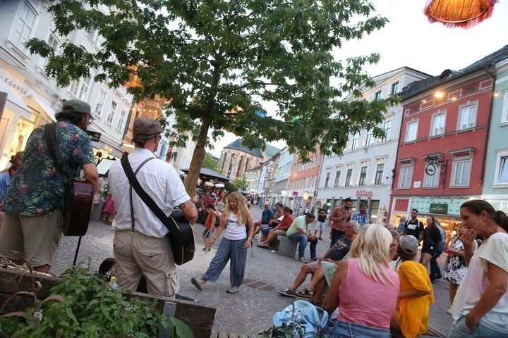 Eine Gruppe von Menschen versammelt sich auf einem Freigelände, möglicherweise einem Stadtplatz, einige sitzen auf Bänken und andere stehen. Zwei Männer spielen Gitarre, einer steht und einer sitzt. Gebäude säumen die Seiten des Platzes.