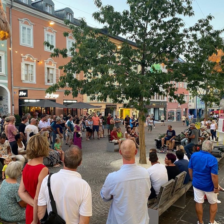 Eine Menschenmenge versammelt sich um einen zentralen Baum auf einem Stadtplatz, einige sitzen auf Bänken und andere stehen. Gebäude säumen die Straße mit Geschäften und Cafés.