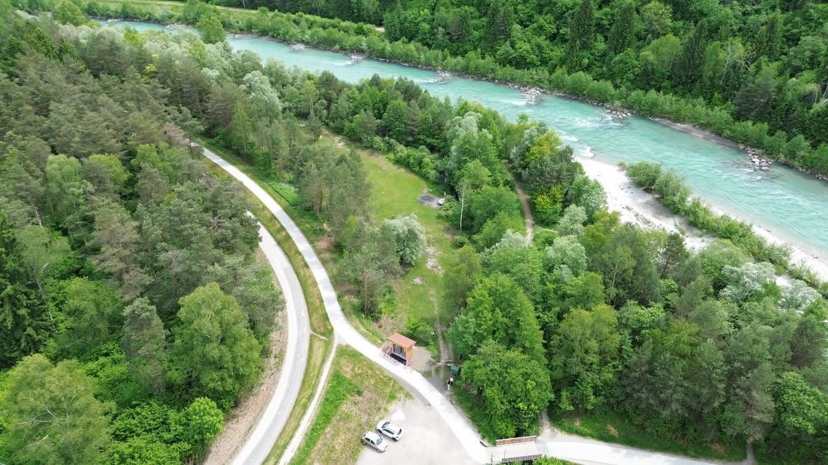 Luftaufnahme eines Flusses mit einer kleinen Hütte und zwei geparkten Autos in der Nähe einer Straße. Der Fluss ist von üppigem grünem Laub und Pflanzen umgeben.