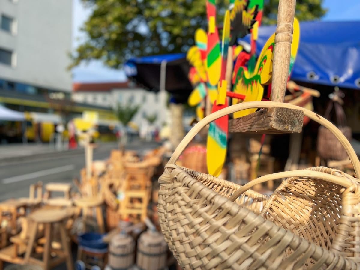 Ein Marktstand mit einem Korb und bunten Holzspielzeug im Vordergrund, mit einem unscharfen Hintergrund von einer Straße und Bäumen.
