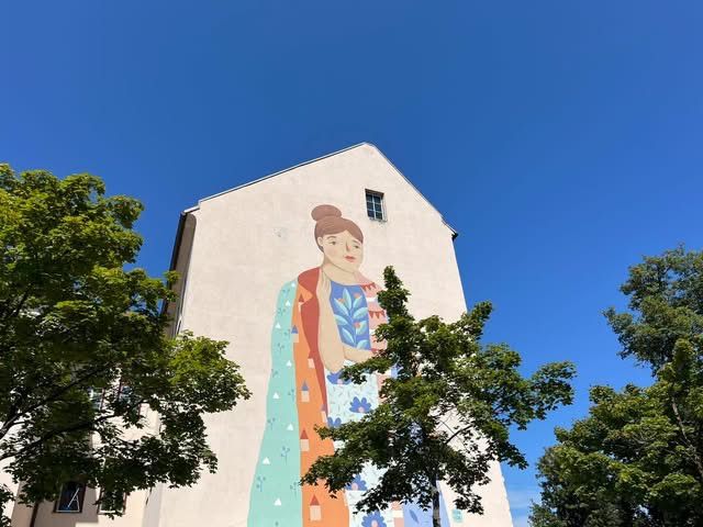 Ein großes Wandbild einer Frau mit Dutt und einem gemusterten Kleid schmückt die Seite eines Gebäudes unter einem klaren blauen Himmel.