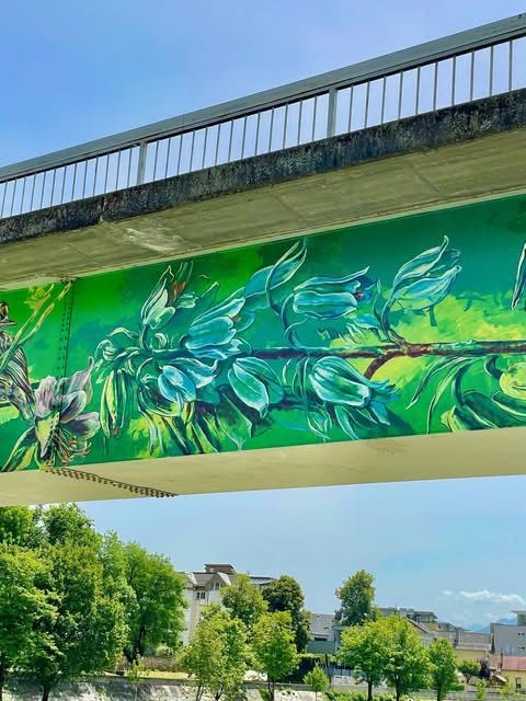 Ein farbenfrohes Wandbild von blauen Blumen und Blättern, das auf der Unterseite einer Brücke mit einem Metallgeländer gemalt ist, vor dem Hintergrund von Bäumen und Gebäuden unter einem klaren Himmel.