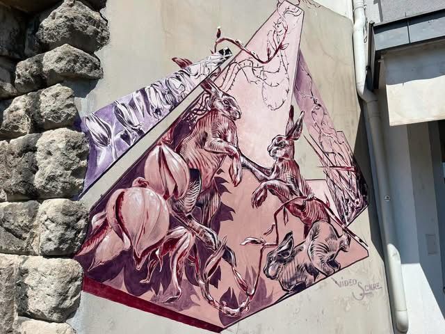 Ein farbenfrohes Wandbild an einer Steinmauer zeigt eine geometrische Komposition von Kaninchen in verschiedenen Posen, mit einer Mischung aus Lila und Rosa Tönen.