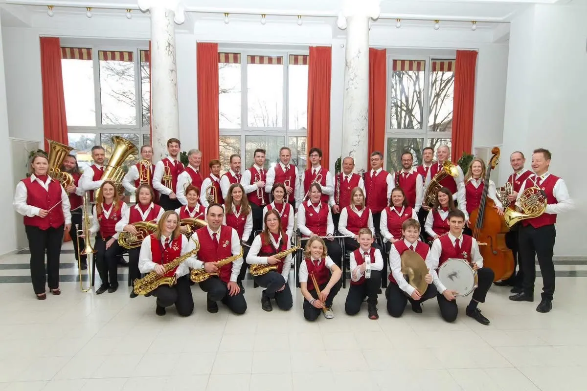 Eine Gruppe von Musikern in roten Westen und weißen Hemden posiert für ein Foto und hält verschiedene Instrumente wie Trompeten, Saxophone und Trommeln. Sie befinden sich in einem Raum mit weißen Wänden, roten Vorhängen und großen Fenstern.
