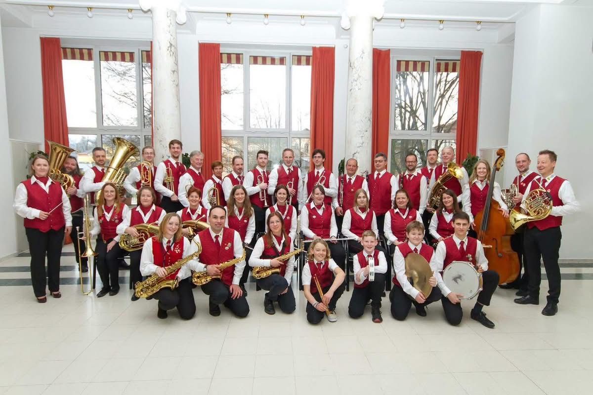 Eine Gruppe von Musikern in roten Westen und weißen Hemden posiert für ein Foto und hält verschiedene Instrumente wie Trompeten, Saxophone und Trommeln. Sie befinden sich in einem Raum mit weißen Wänden, roten Vorhängen und großen Fenstern.