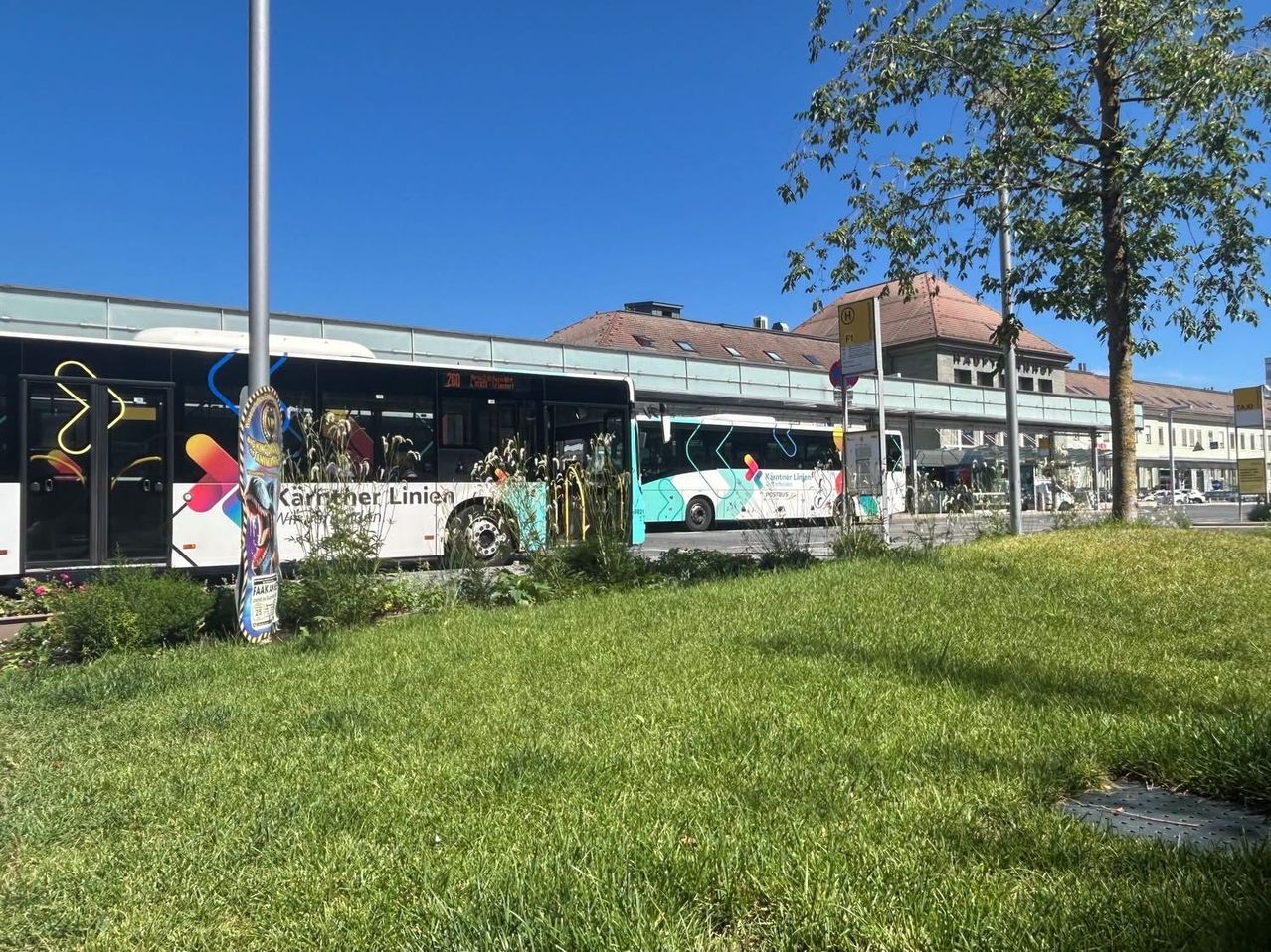 Ein Busbahnhof mit zwei geparkten Bussen, einer mit bunten Designs und der andere mit einem weißen und blauen Farbschema. Der Bahnhof ist von einer Rasenfläche und einem Gebäude mit einem braunen Dach umgeben.