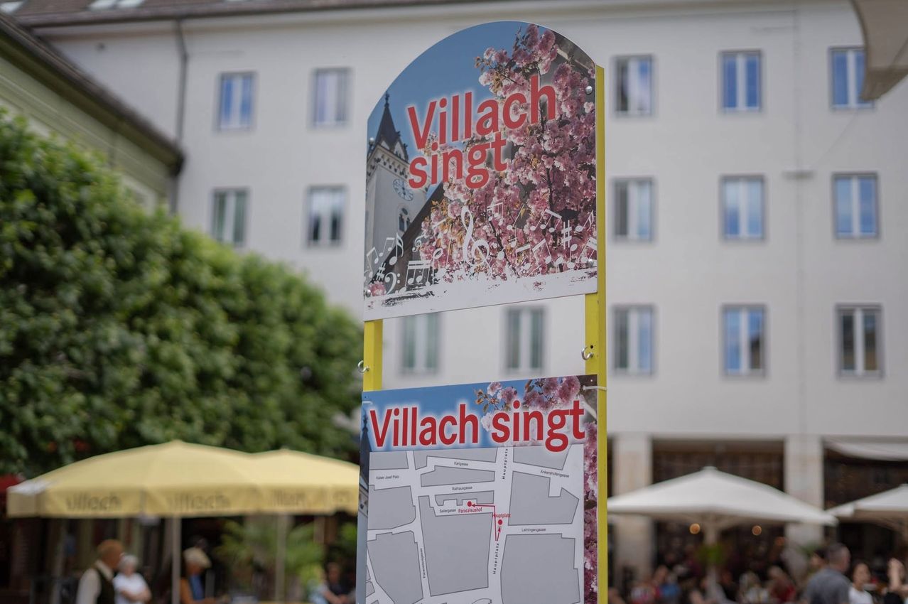 A signboard with 'Villach singst' in red on a yellow pole, showcasing a map and cherry blossoms against a white building.