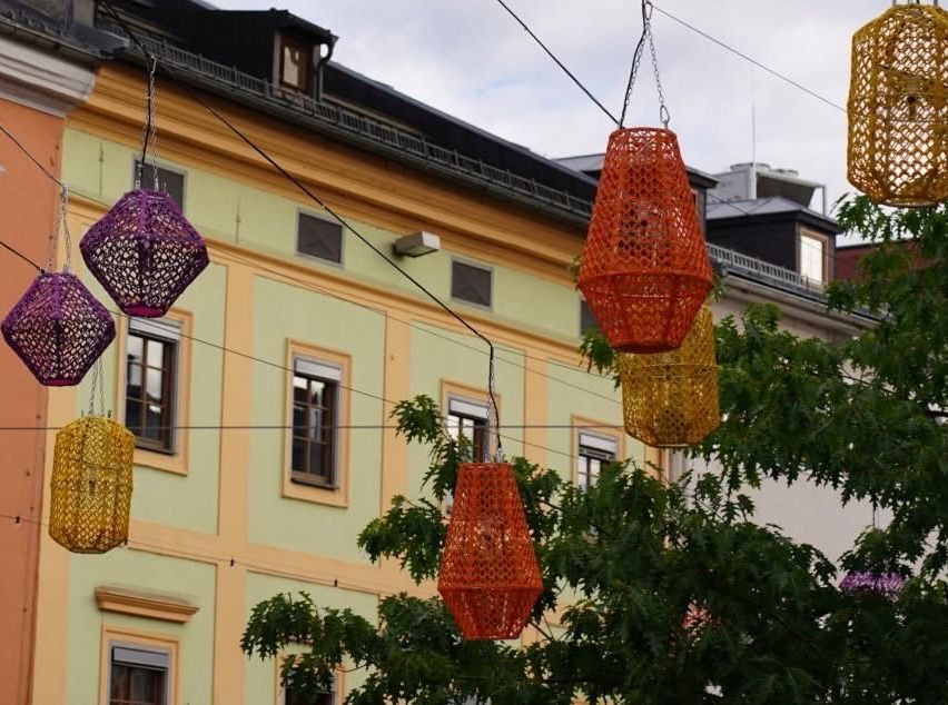 Colorful knitted lanterns hang from wires above a street, with a tree and buildings in the background.