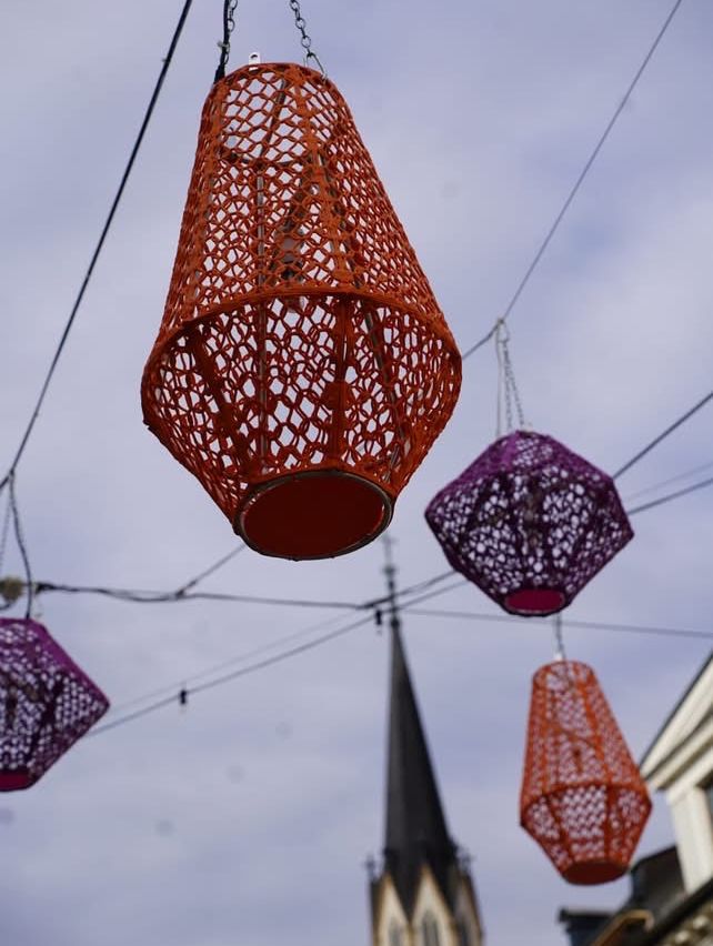 Four colorful, netted lanterns are hanging from wires against a cloudy sky with a church spire in the background.