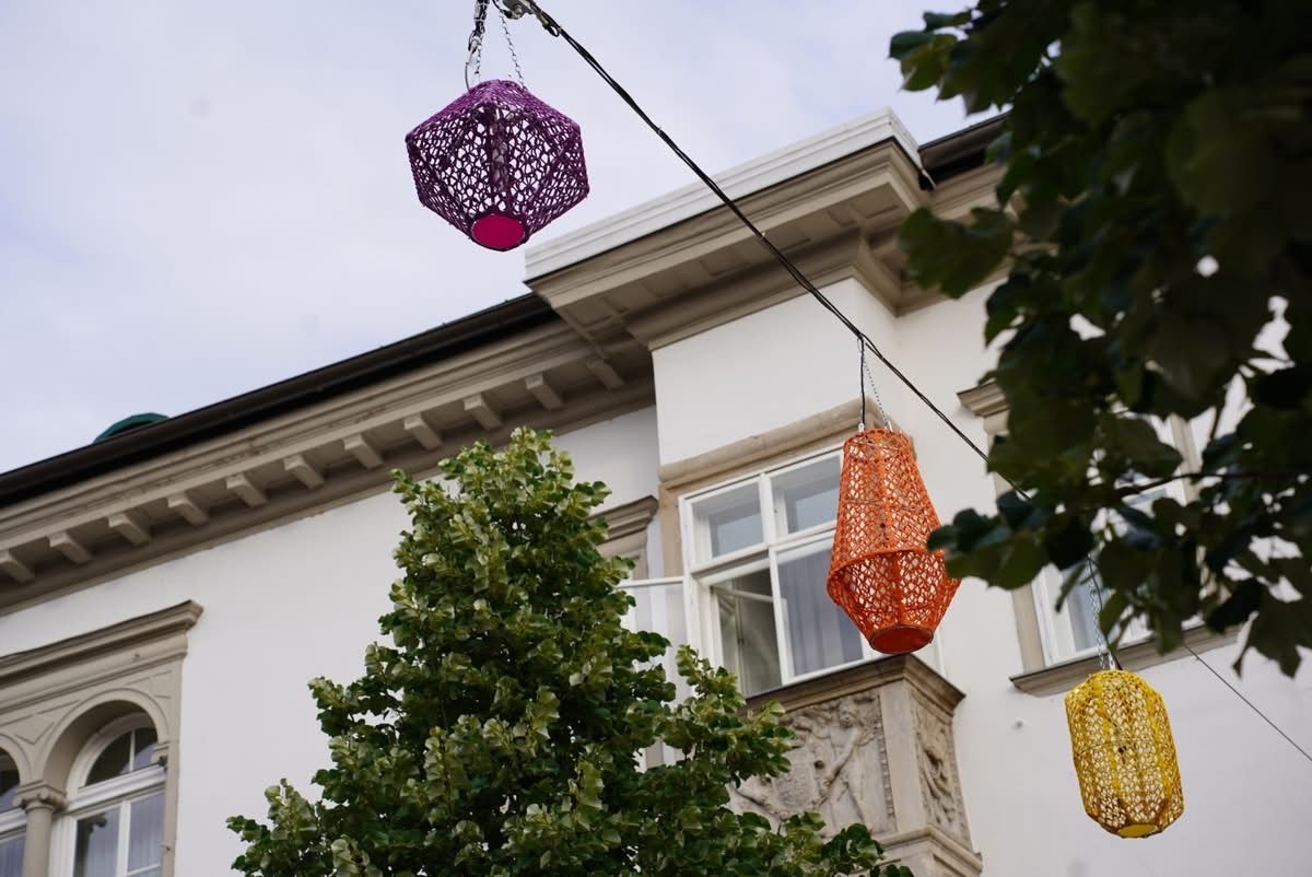 A tree with a decorative purple lantern hangs from a wire above a white building with an ornate balcony. An orange lantern hangs from a wire on the right.