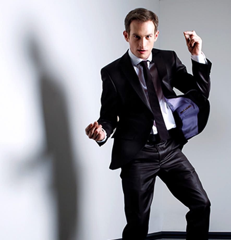 A man in a black suit is posing with his jacket in his hand, standing against a light-colored wall.