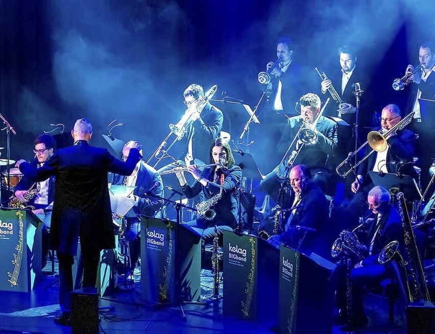 A jazz band is performing on stage with a blue background, wearing formal attire. A man in front is standing and conducting.