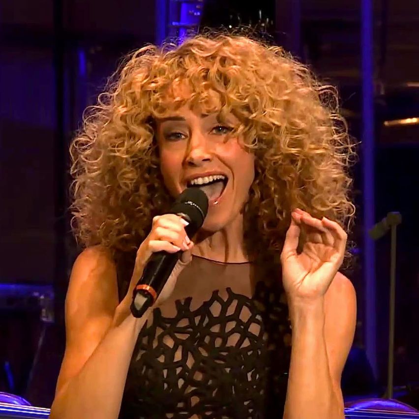 A woman with curly blonde hair sings into a microphone on stage, smiling, with hands raised. The background is dark with blue lighting.