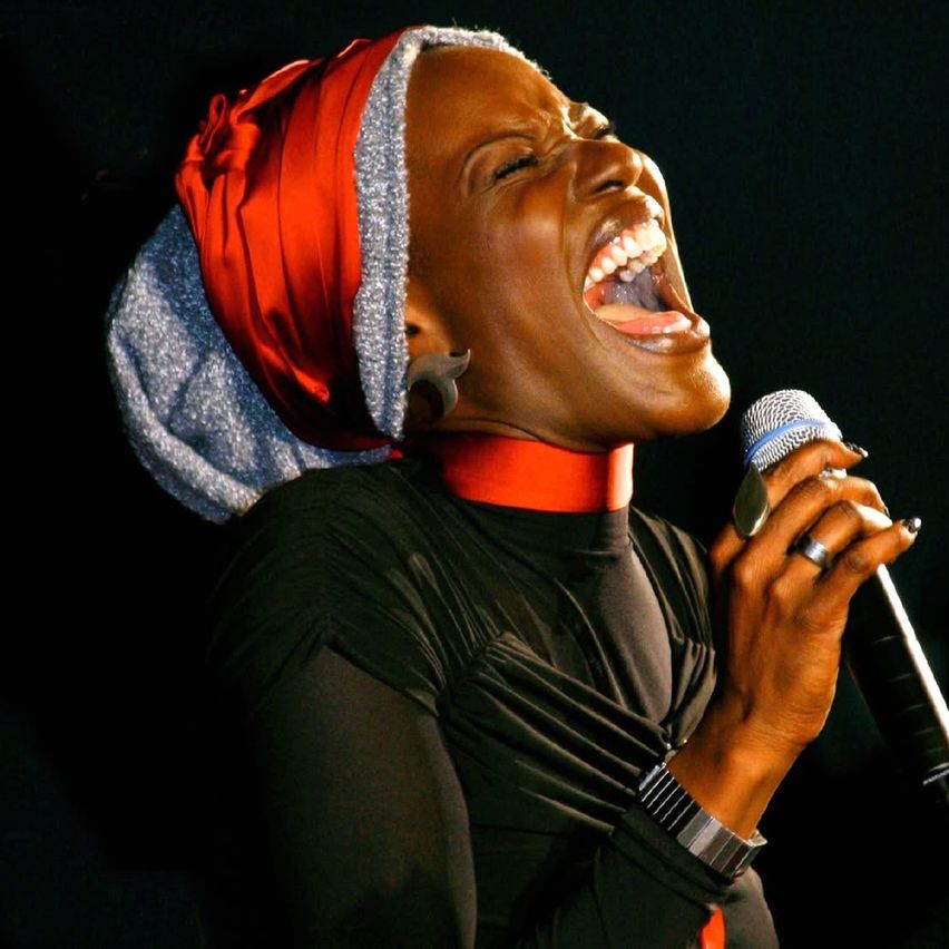 A woman sings on stage with a microphone, wearing a black shirt, a red scarf, and a ring. Her eyes are closed and her mouth is open.