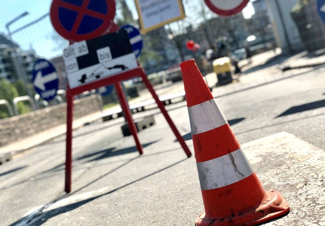A traffic cone stands in front of a red signboard with 'Gilt ab' and 'bis' written on it. Other signs are blurry in the background.