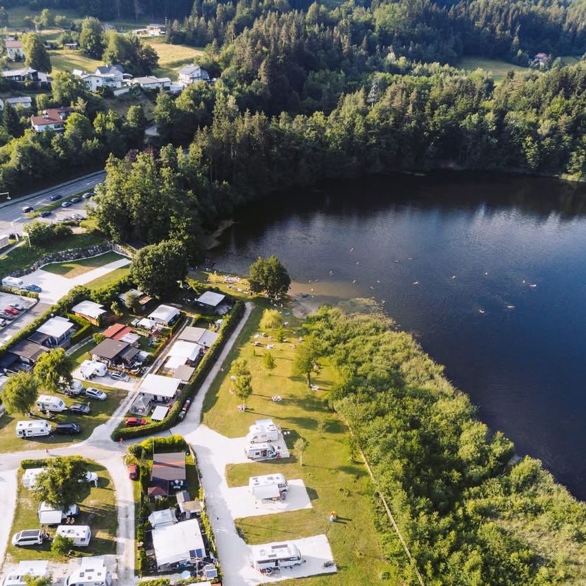 Aerial view of a campsite beside a lake with several tents and recreational areas. Trees and houses are visible in the background.