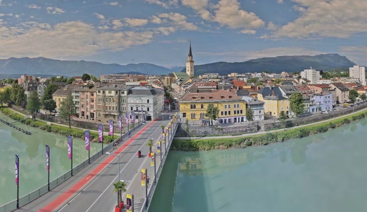 Luftaufnahme einer Stadt mit einer Brücke über einem Fluss, Gebäuden und einem Turm in der Ferne unter einem bewölkten Himmel.