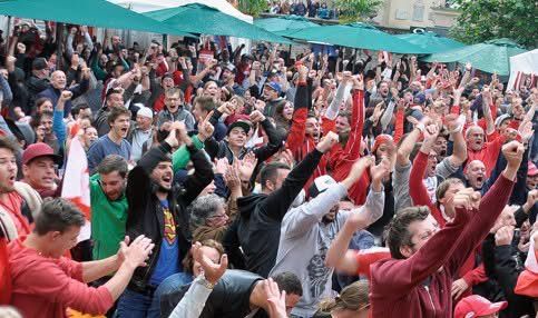 Eine Menschenmenge feiert im Freien unter grünen Schirmen, viele heben die Hände und tragen rote Shirts.