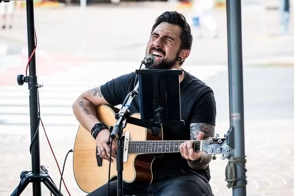 Ein Mann spielt Gitarre und singt in ein Mikrofon auf der Straße. Er hat Tattoos und trägt ein schwarzes Shirt.