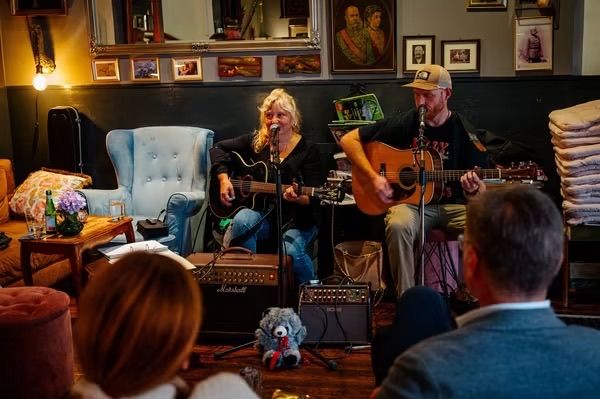 Zwei Personen spielen Gitarre und singen in einer gemütlichen Lounge-Atmosphäre, umgeben von einem kleinen Publikum.