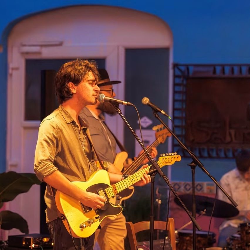 Zwei Männer spielen auf der Bühne bei Nacht, einer singt in ein Mikrofon, während er eine gelbe Gitarre spielt. Ein anderer Mann spielt Gitarre hinter ihm, mit Trommeln und Becken im Hintergrund.