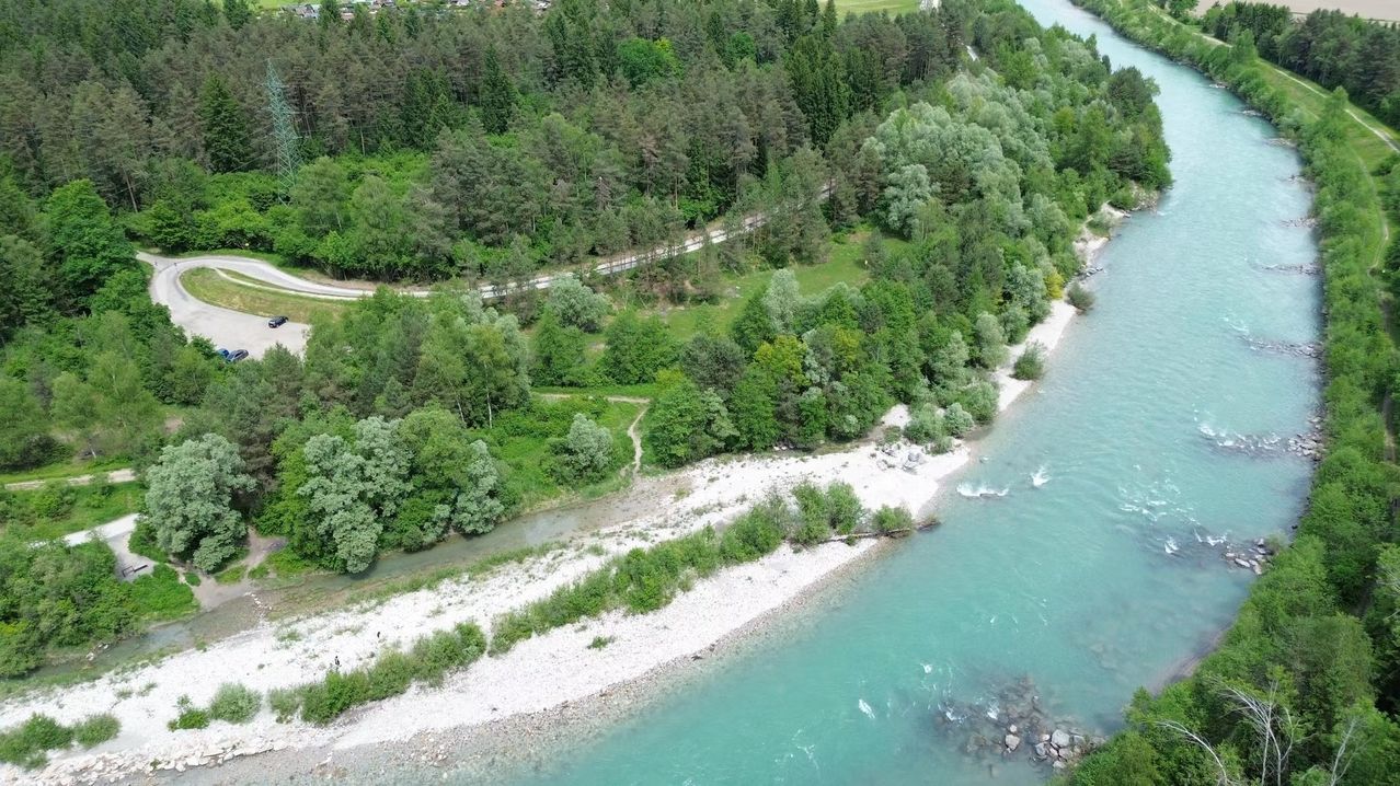 Luftaufnahme eines Flusses, der durch einen üppigen Wald fließt. Bäume und Sträucher säumen die Ufer. Ein Weg führt vom Fluss zu einer entfernten Straße.