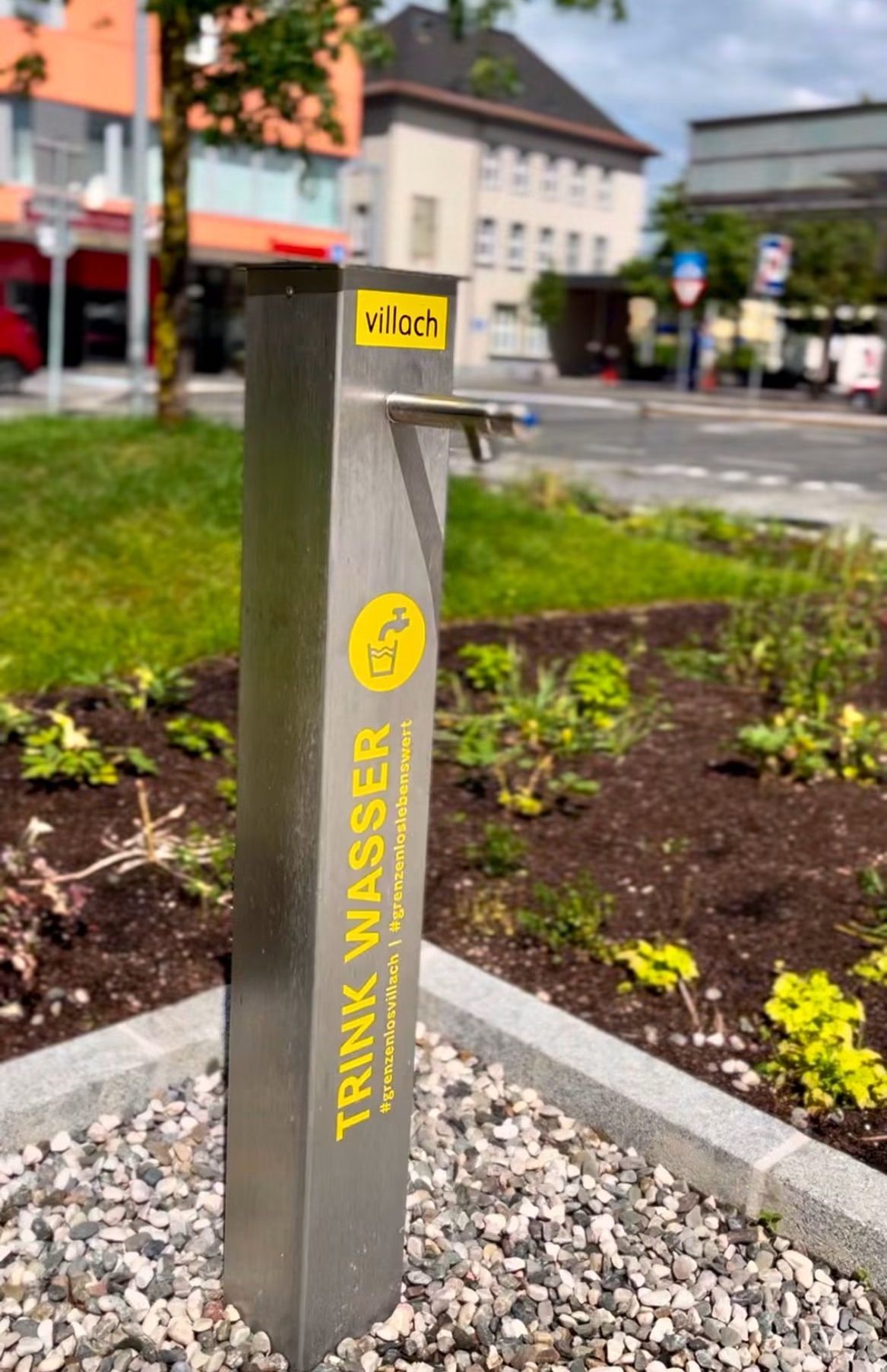 Ein metallischer Wasserbrunnen mit gelbem Text auf grünem Hintergrund. Es steht 'Trink Wasser' und befindet sich in einem städtischen Bereich.