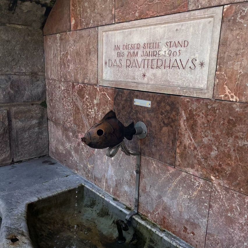 Eine Marmorwand hat einen Wasserhahn mit Fischdesign. Ein Waschbecken befindet sich unter dem Wasserhahn. Über dem Wasserhahn befindet sich eine Tafel mit Text. Der Text ist in Deutsch geschrieben.